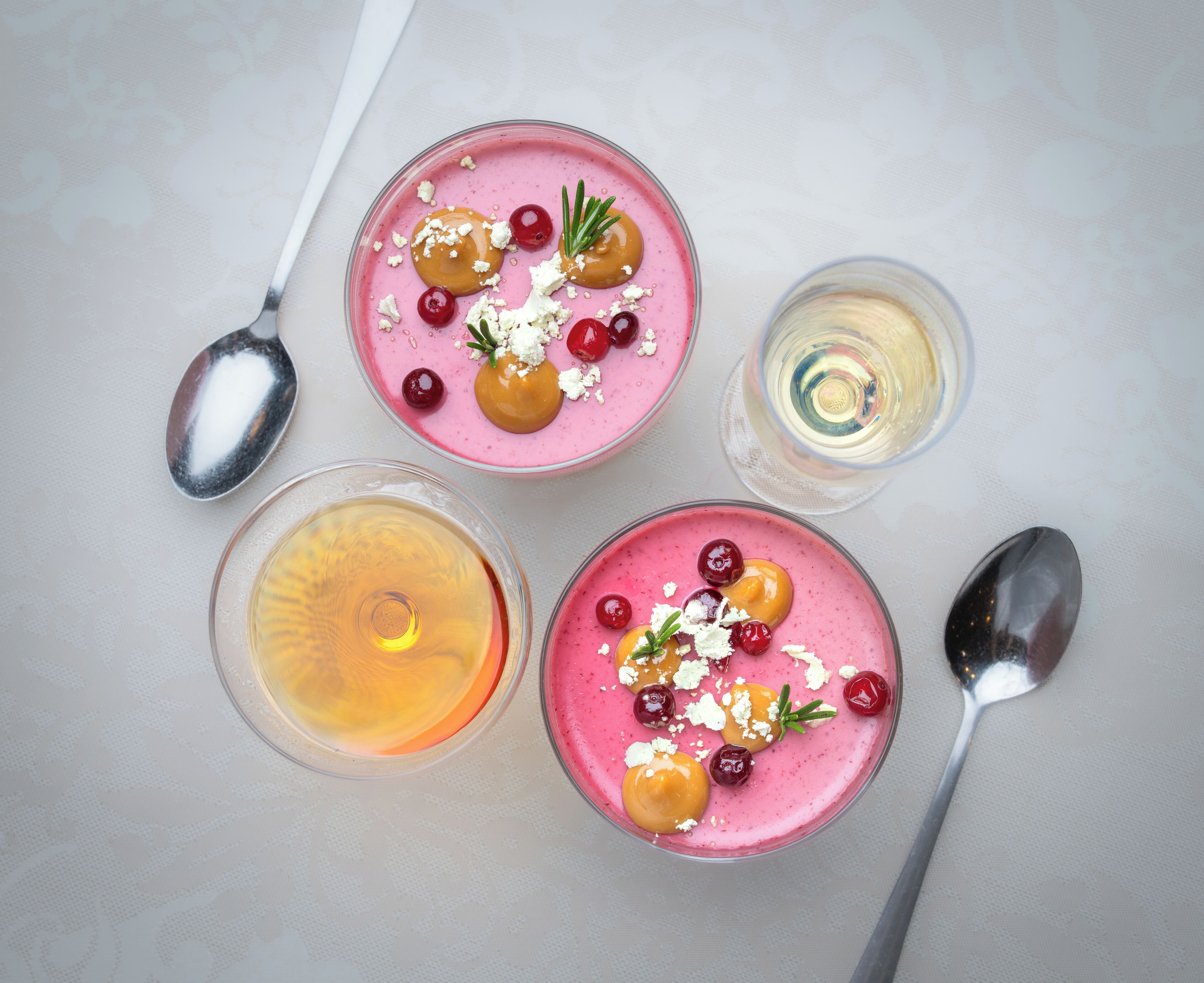 Vier glazen roze smoothies met toppings, twee lepels, en een klein glas witte vloeistof.