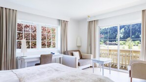 A hotel room with a large bed, a desk, a chair, a sofa, and a balcony with a view of trees and a building. - Loenfjord (Stryn)