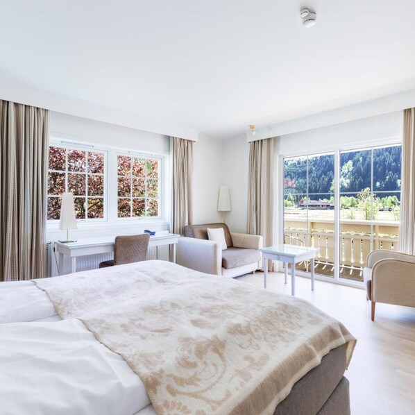 Hotelkamer met een groot bed, een bureau, een stoel, een bank en een balkon met uitzicht op bomen en een gebouw.