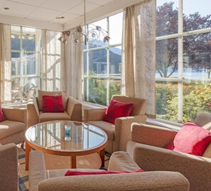 Een kamer met een glazen salontafel, beige banken met rode kussens en grote ramen met uitzicht op bomen en bergen.
