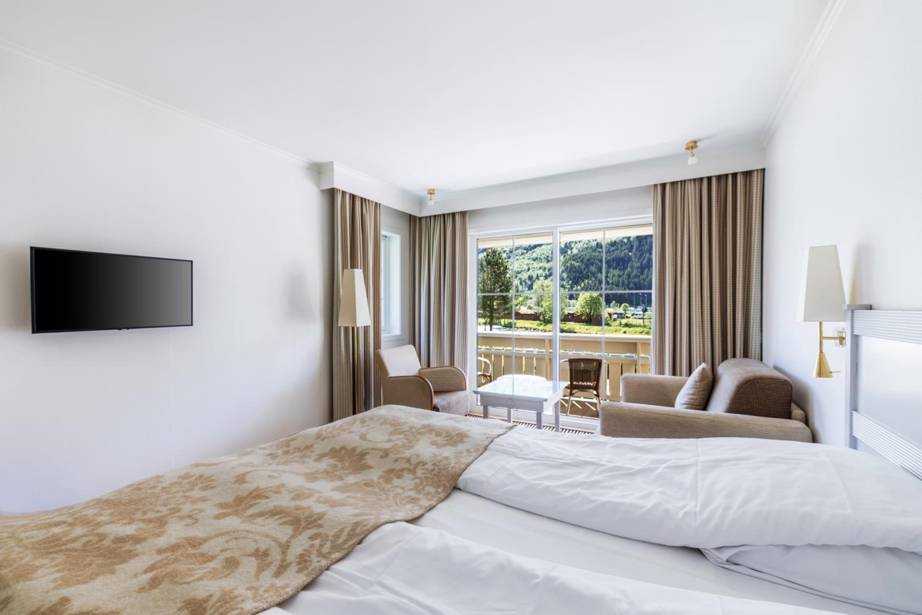 Una habitación de hotel con cama, televisión, balcón con mesa y sillas, y cortinas.