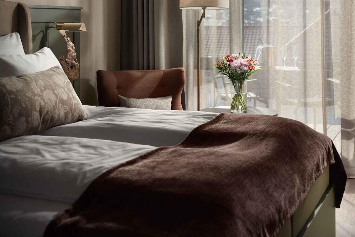 A hotel room with a bed, a chair, a lamp, and a view of the city through large windows.