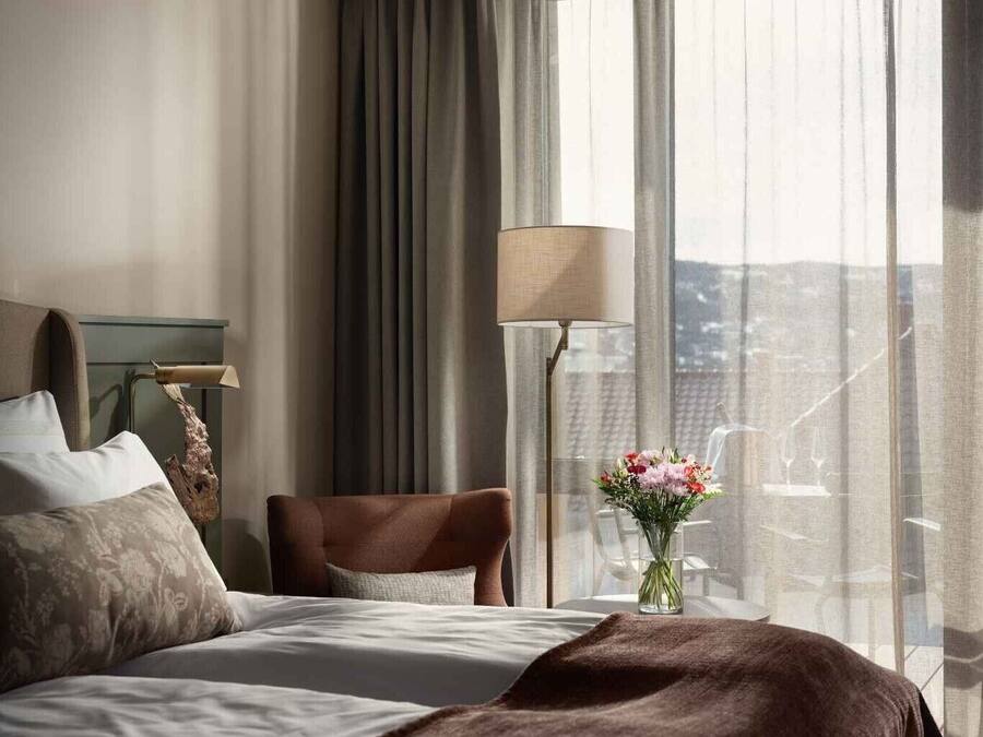A hotel room with a bed, a chair, a lamp, and a view of the city through large windows.
