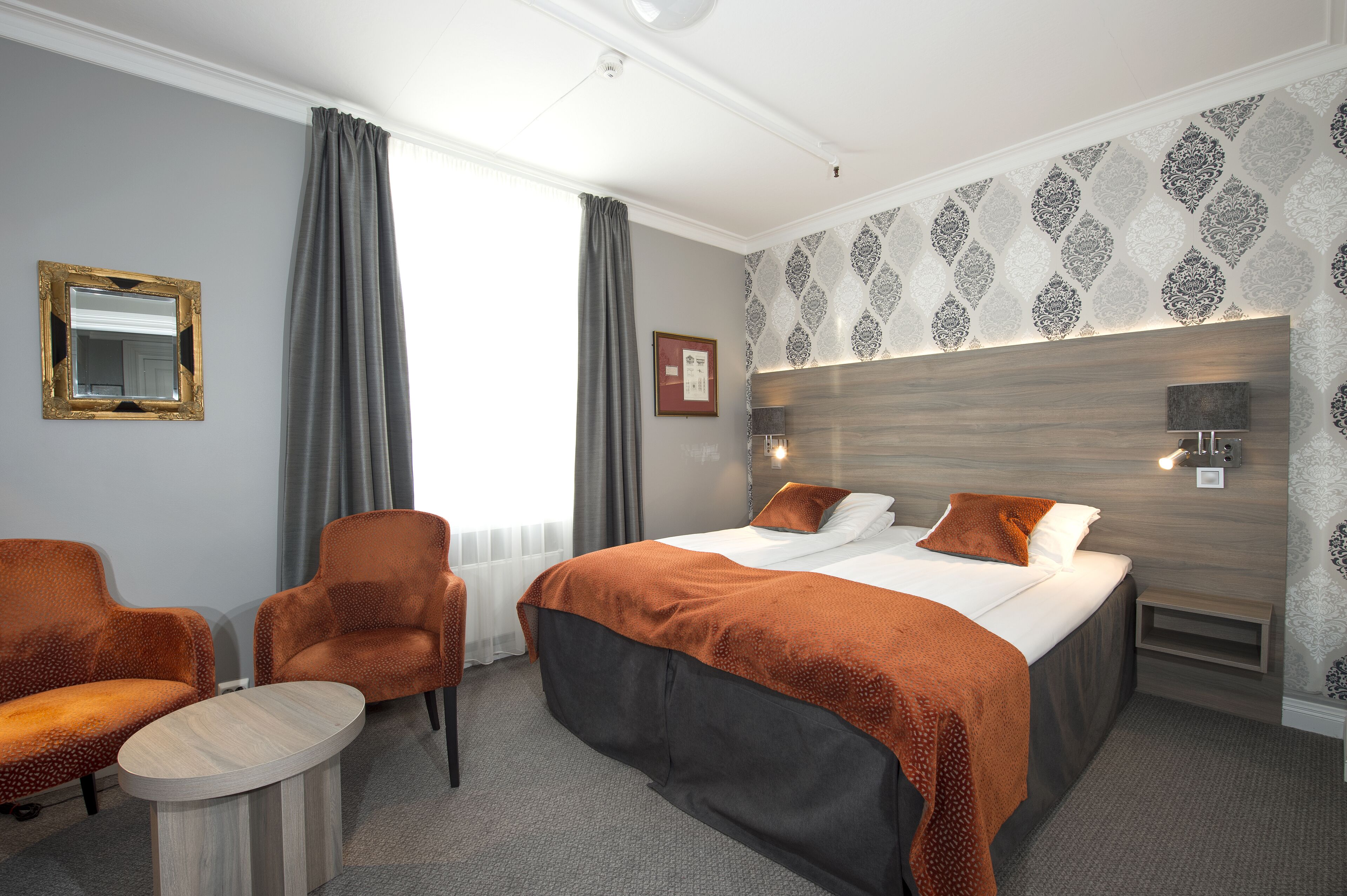 Une chambre d’hôtel avec un lit, deux chaises, une petite table, un miroir et un mur décoré de motifs.