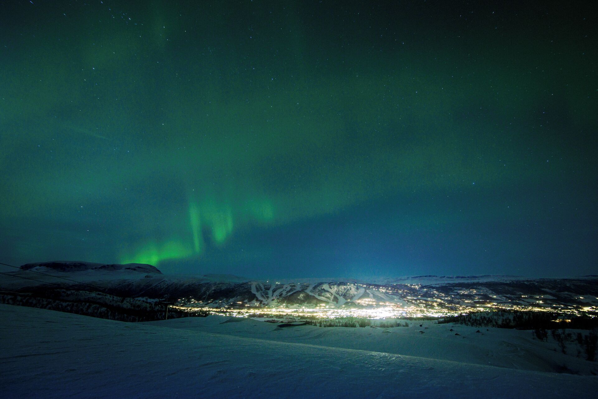 Un paysage enneigé avec une ville illuminée au loin, sous un ciel nocturne où se déploie l’aurore boréale.