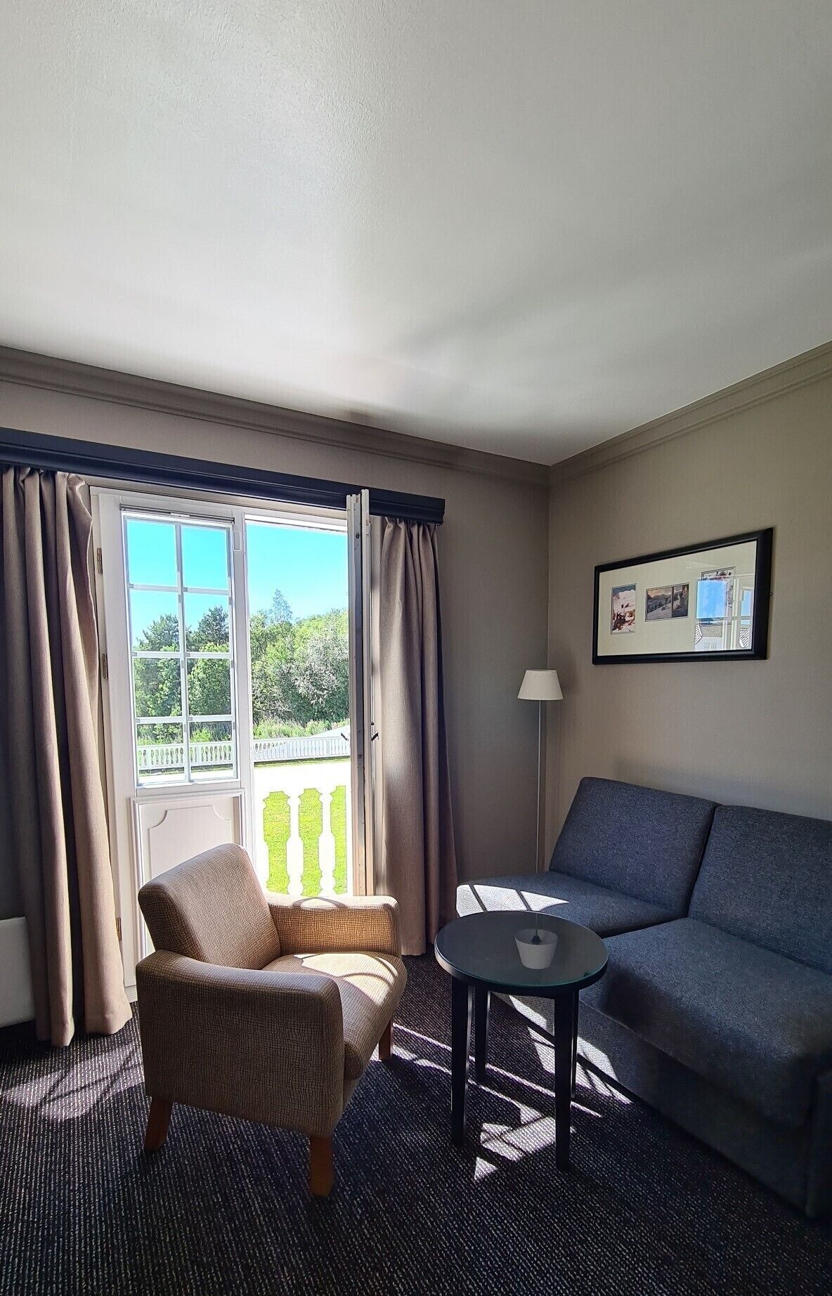 A hotel room with a view of greenery, a sofa, a chair, a small table, and a lamp.