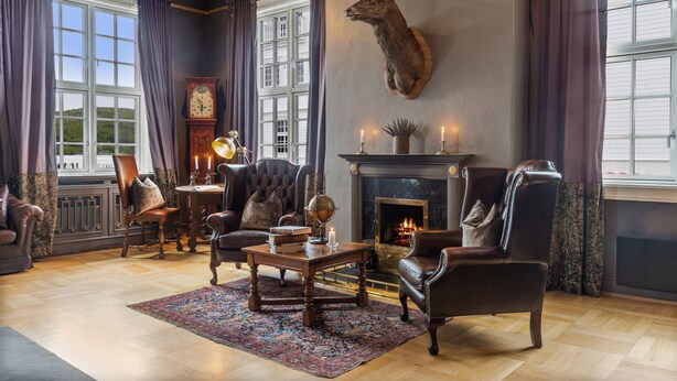 A traditional living room with leather armchairs, a fireplace, a mounted deer head, and a large window with curtains. - Dr. Holms Hotel (Hol)