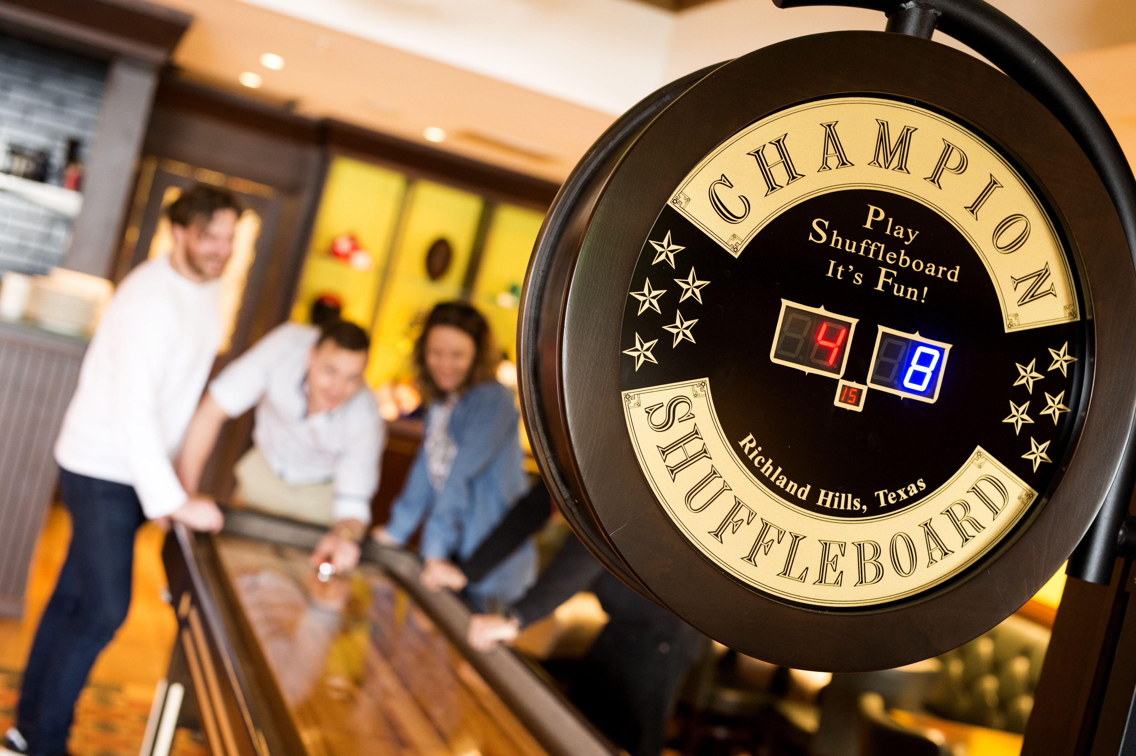 Mesa de shuffleboard com as palavras "CHAMPION" e "SHUFFLEBOARD" visíveis, indicando uma área de jogo temática ou para competição.