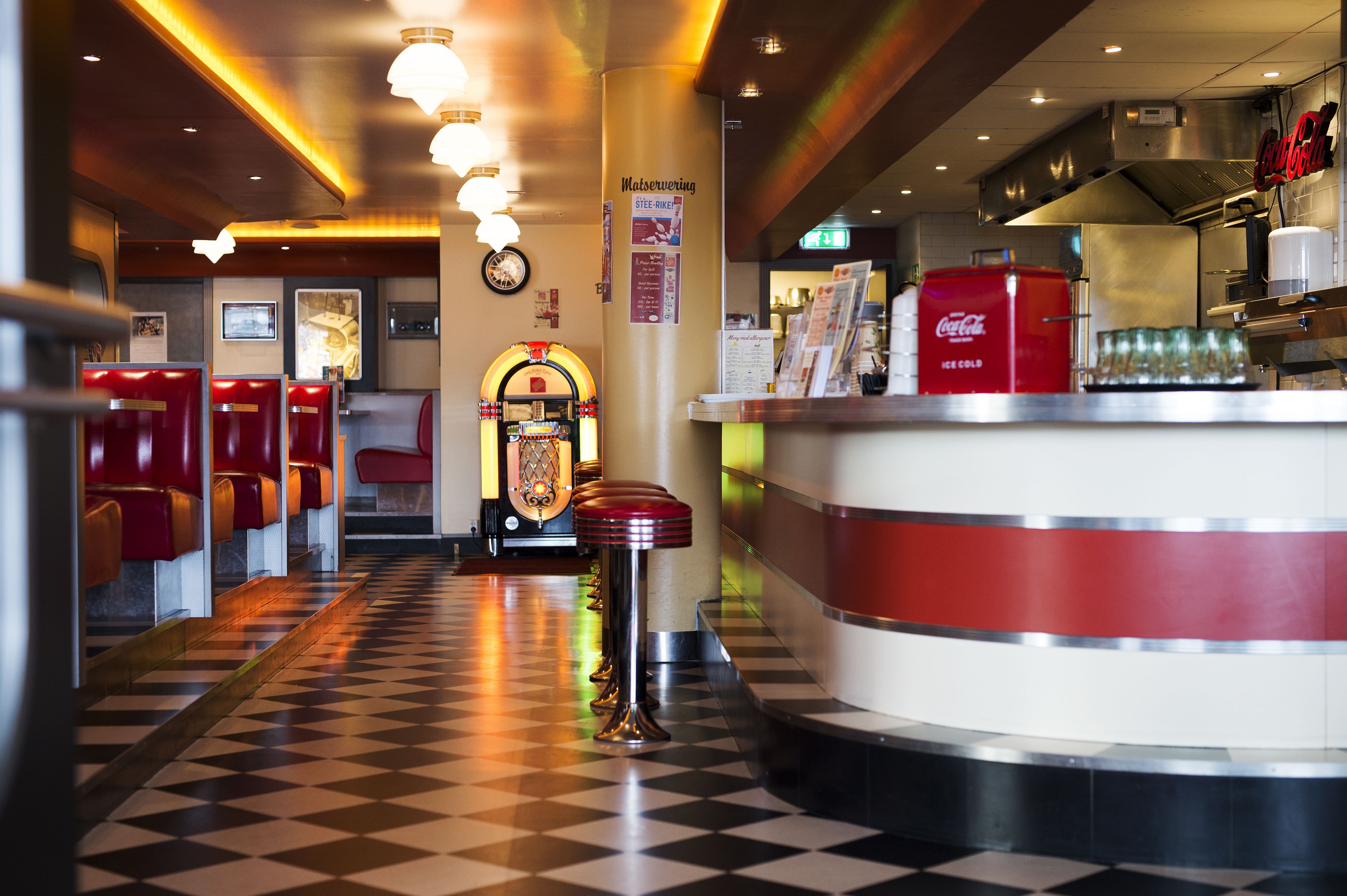 A diner with checkered flooring, red booths, a jukebox, and a Coca-Cola refrigerator.