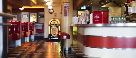 A diner with checkered flooring, red booths, a jukebox, and a Coca-Cola refrigerator.