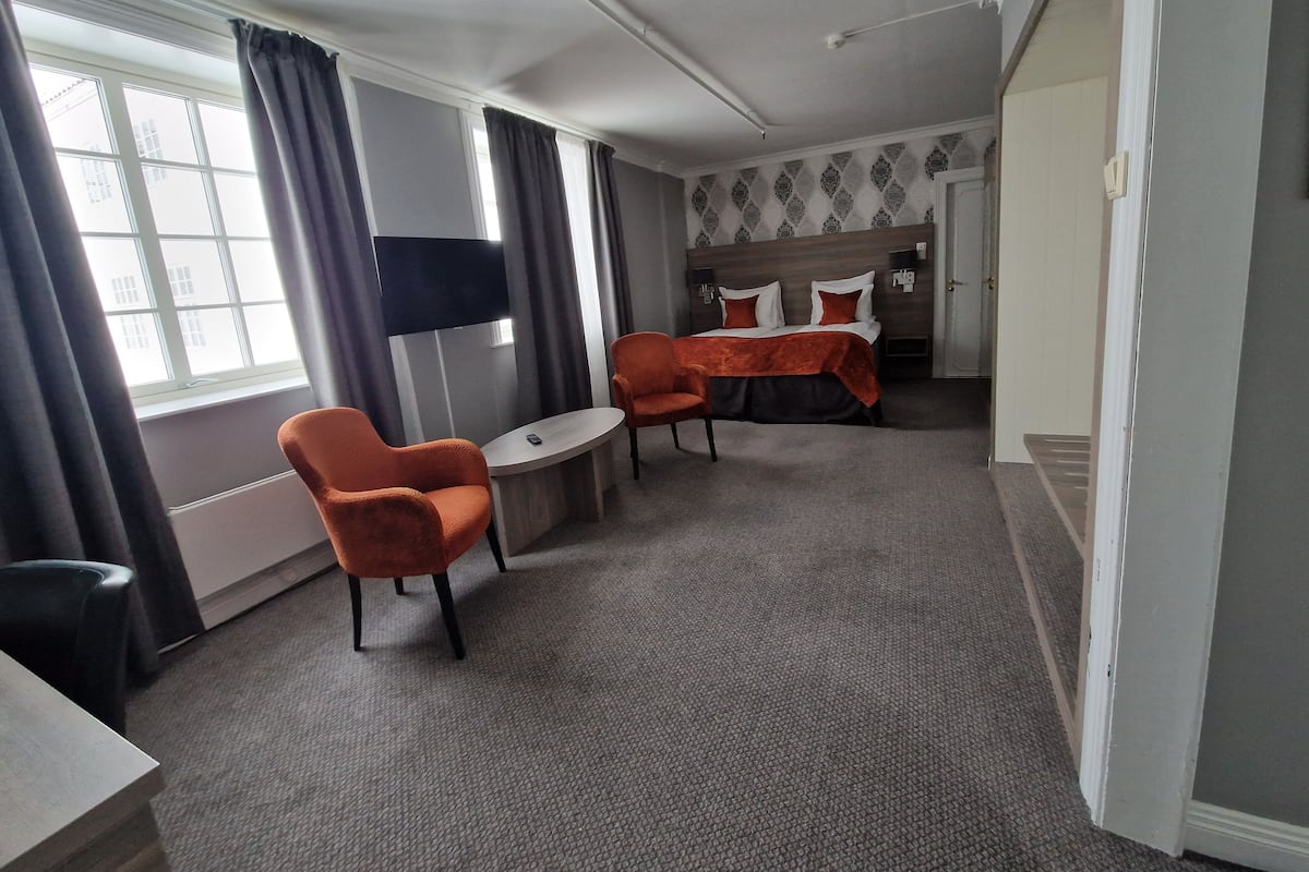 A hotel room with a bed, two orange chairs, a small table, a TV, and a window with curtains.