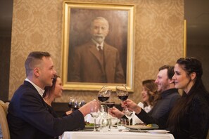 A group of people seated at a table with wine glasses, a framed portrait on the wall, and a patterned wallpaper background.
