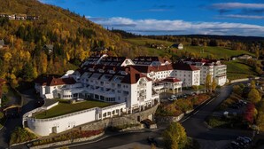 A large, white building with multiple red roofs, surrounded by a parking area and a road. - Dr. Holms Hotel (Hol)