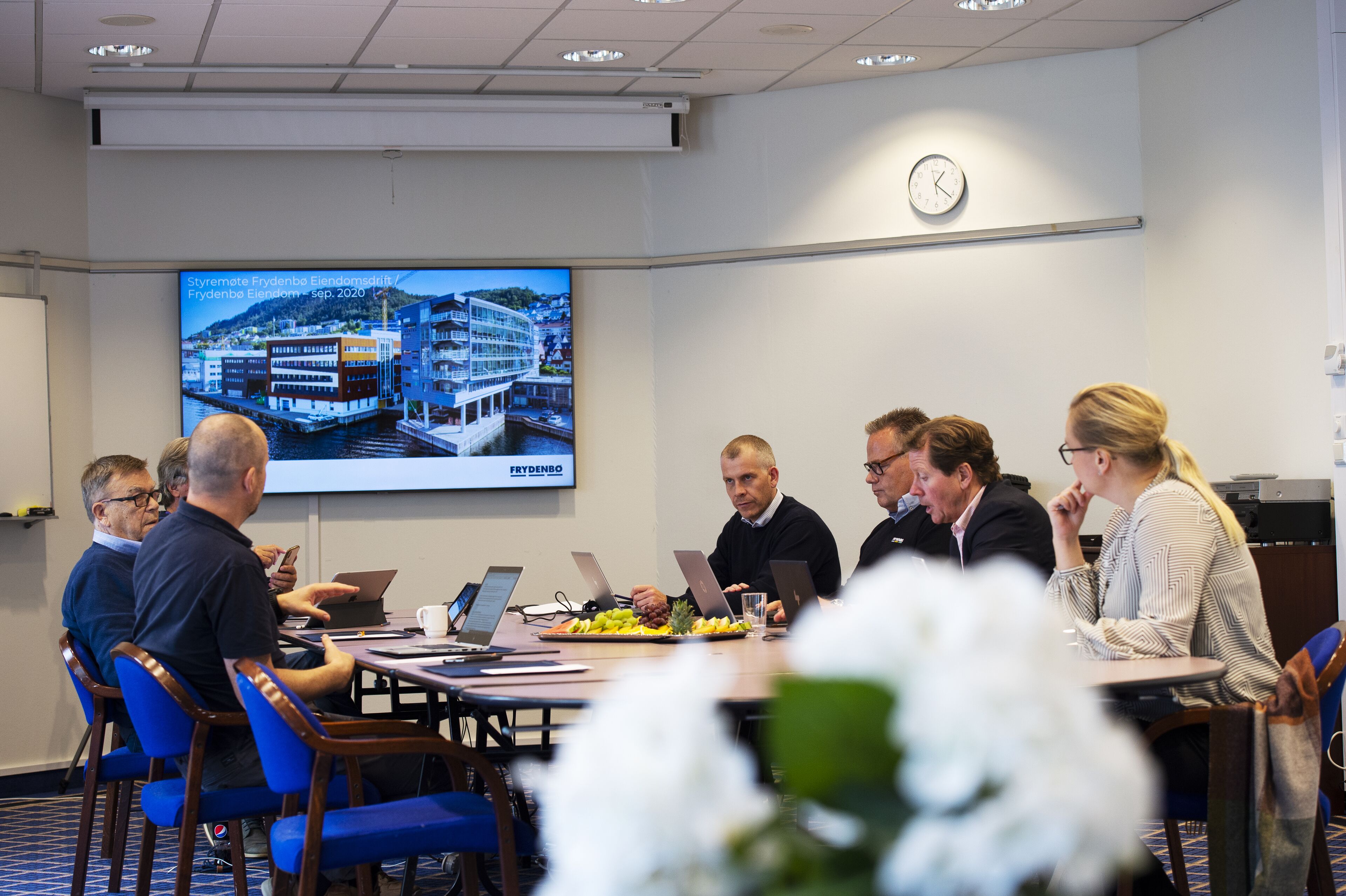 Une salle de conférence équipée d’un écran de présentation, d’une horloge et d’une table sur laquelle sont disposés des ordinateurs portables et une assiette de fruits.