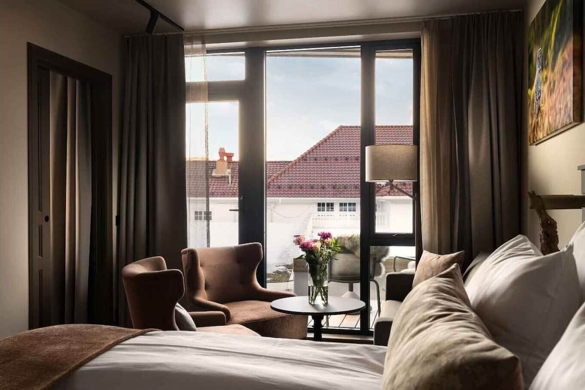 A hotel room with a large bed, a seating area with chairs, a small table with a vase, and a view of buildings outside.