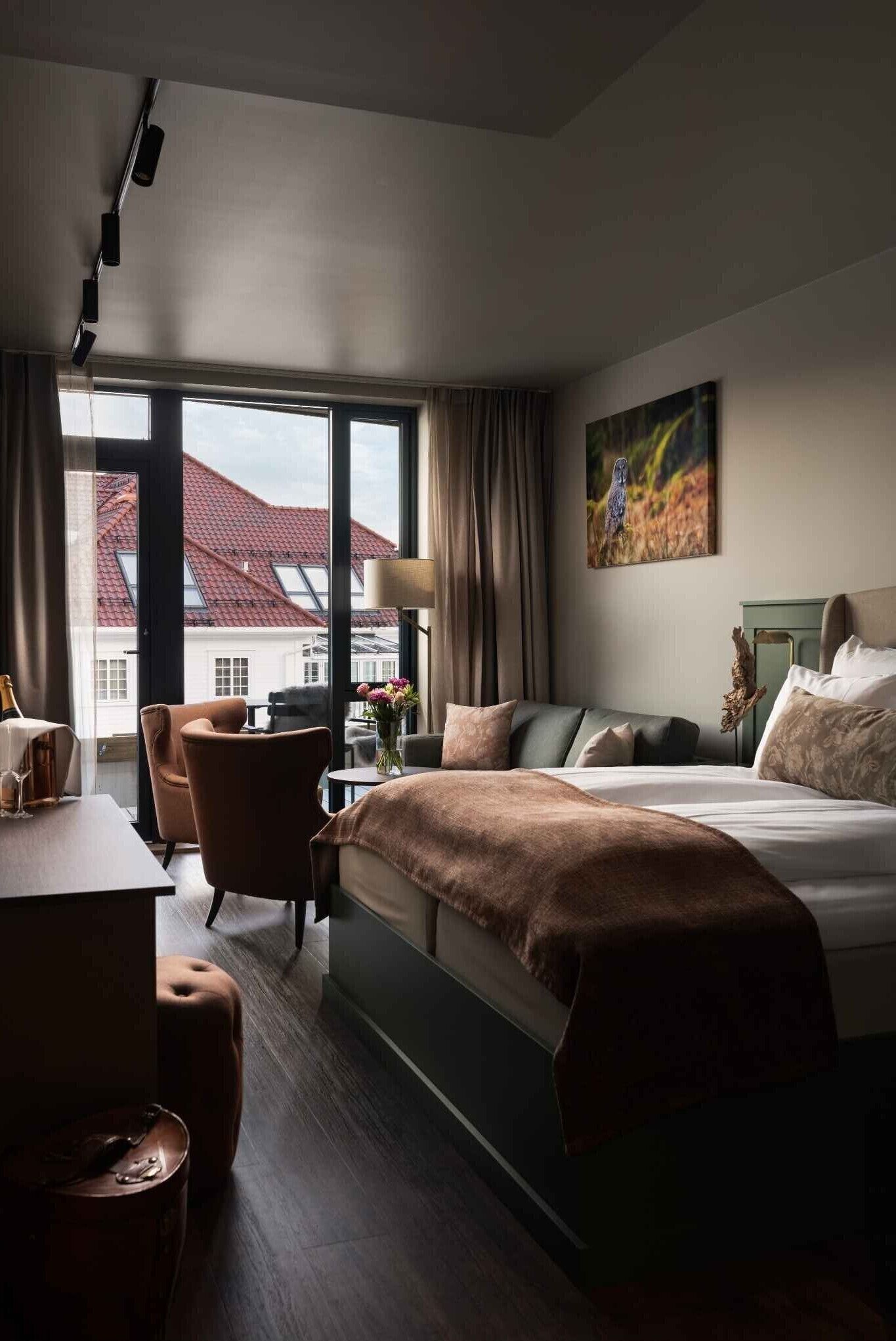 A hotel room with a bed, a desk, a chair, and a large window offering a view of buildings outside.