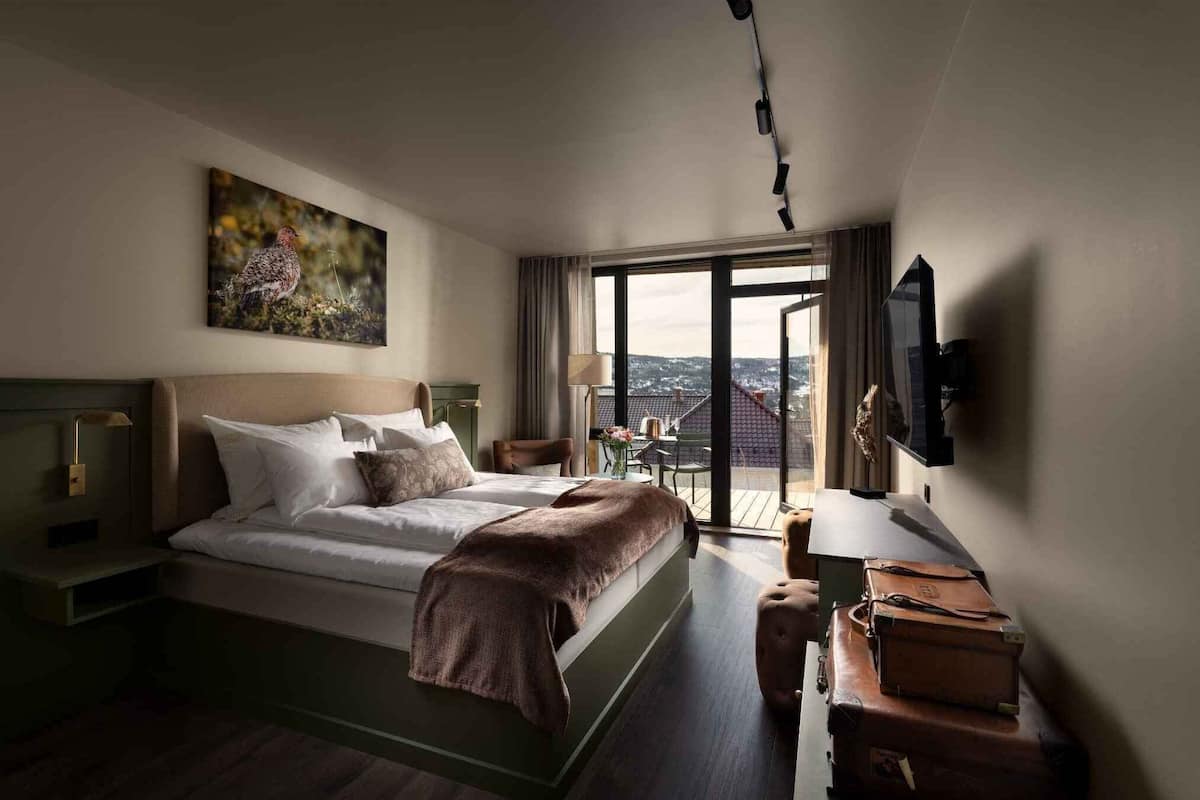 A hotel room with a bed, a television, a desk, a chair, a window with a view, and a painting on the wall.