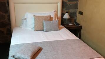 A neatly made bed with a white headboard, a grey throw blanket, and several pillows. A nightstand with a lamp and a phone is beside the bed. The room has a stone wall and a plain wall.