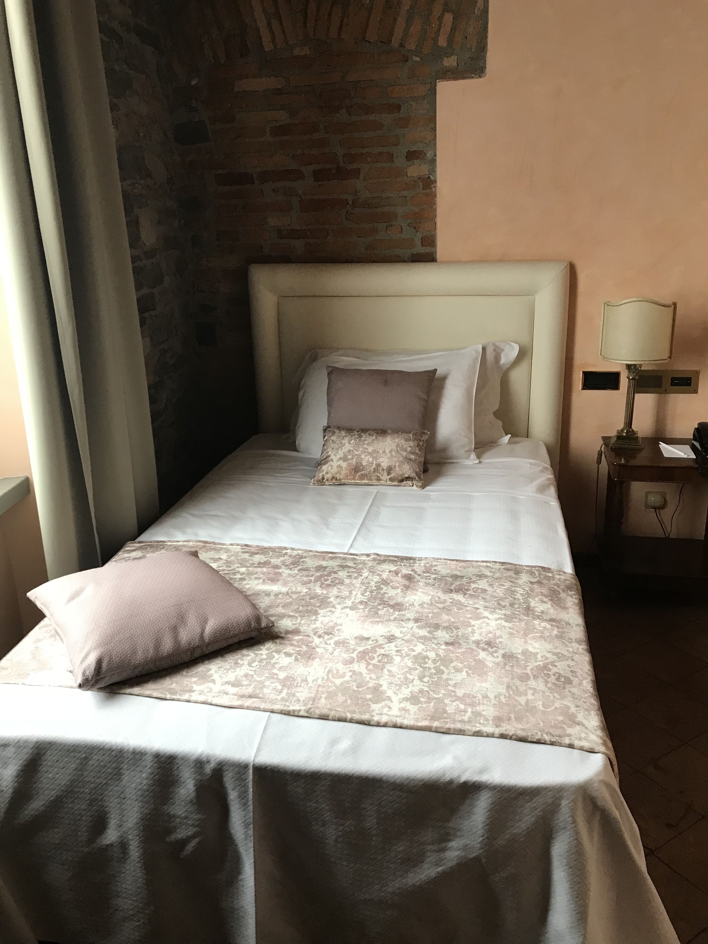 Una cama bien hecha con una colcha estampada, una almohada y un cabecero frente a una pared de ladrillo.