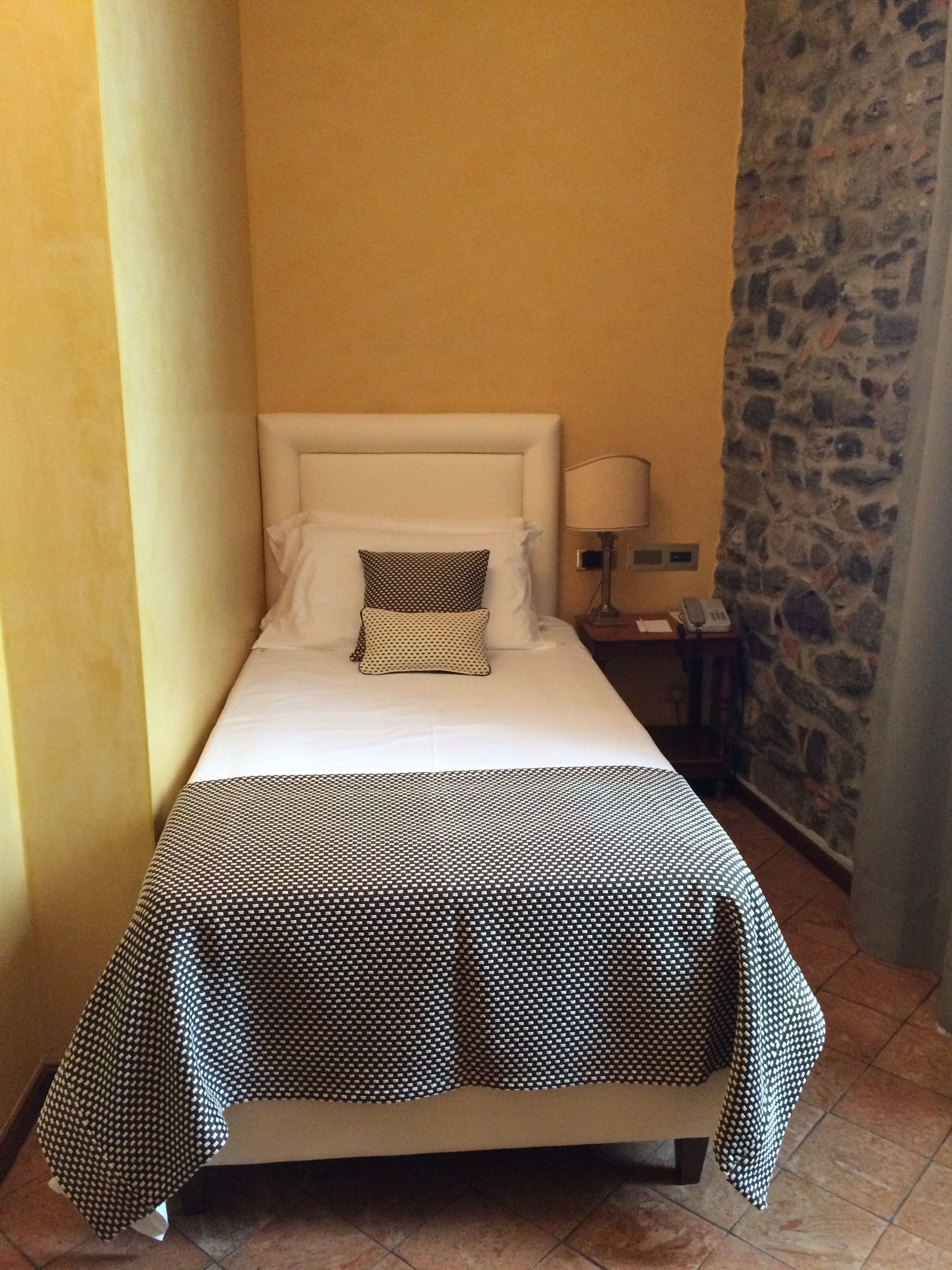 A small, single-bed room with a white headboard, a patterned bedspread, and a bedside table with a lamp.