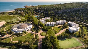 Eine Luftaufnahme eines Resorts mit mehreren Gebäuden, einer zentralen Kuppelstruktur, einem Tennisplatz und einem Strand.