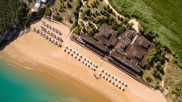 Ein Luftbild eines Hotels direkt am Strand mit einer langen Reihe von Strandsonnenschirmen und einem Gebäude mit flachem Dach.