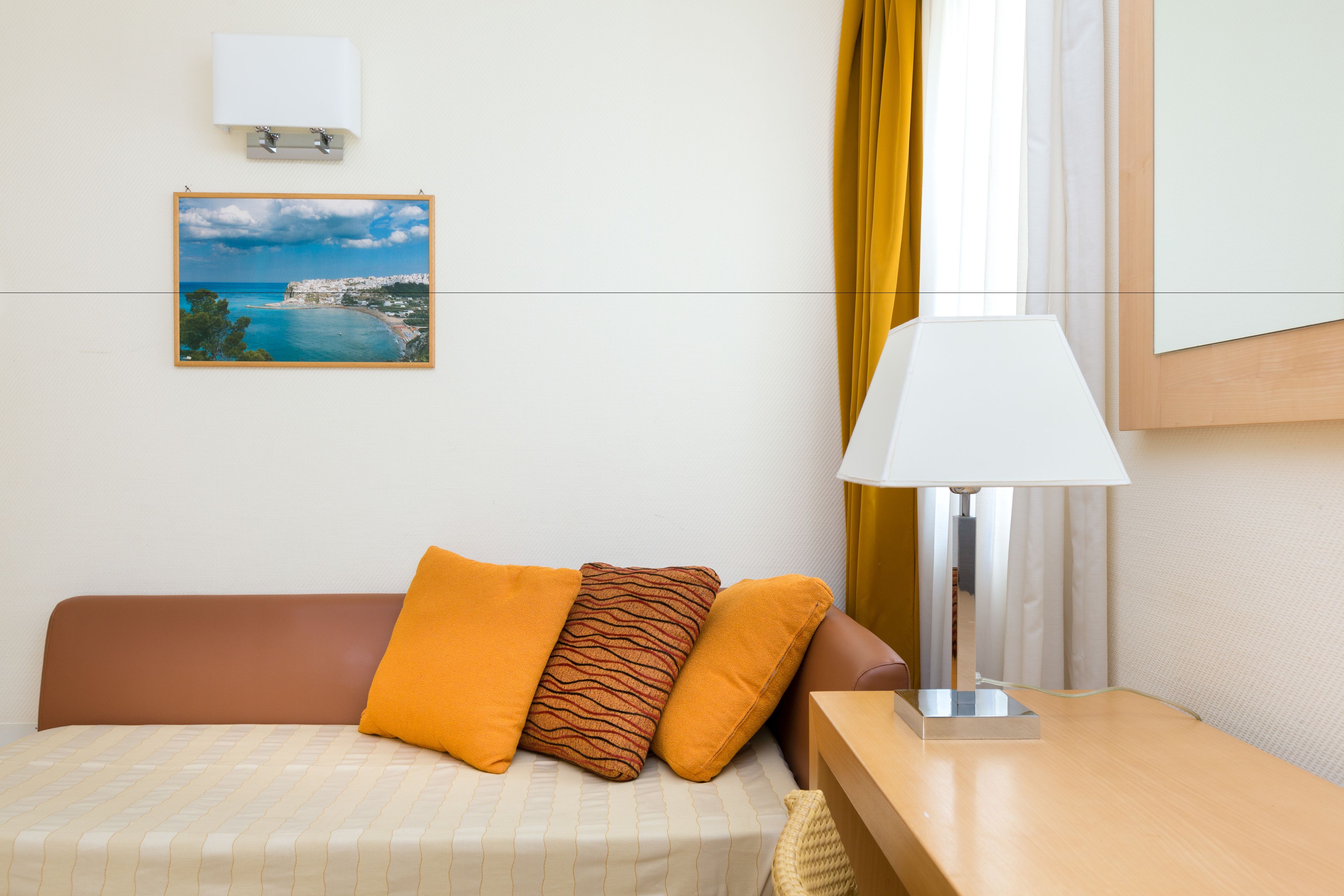 A hotel room with a bed, a bedside lamp, a framed picture of a coastal landscape, and a window with curtains.