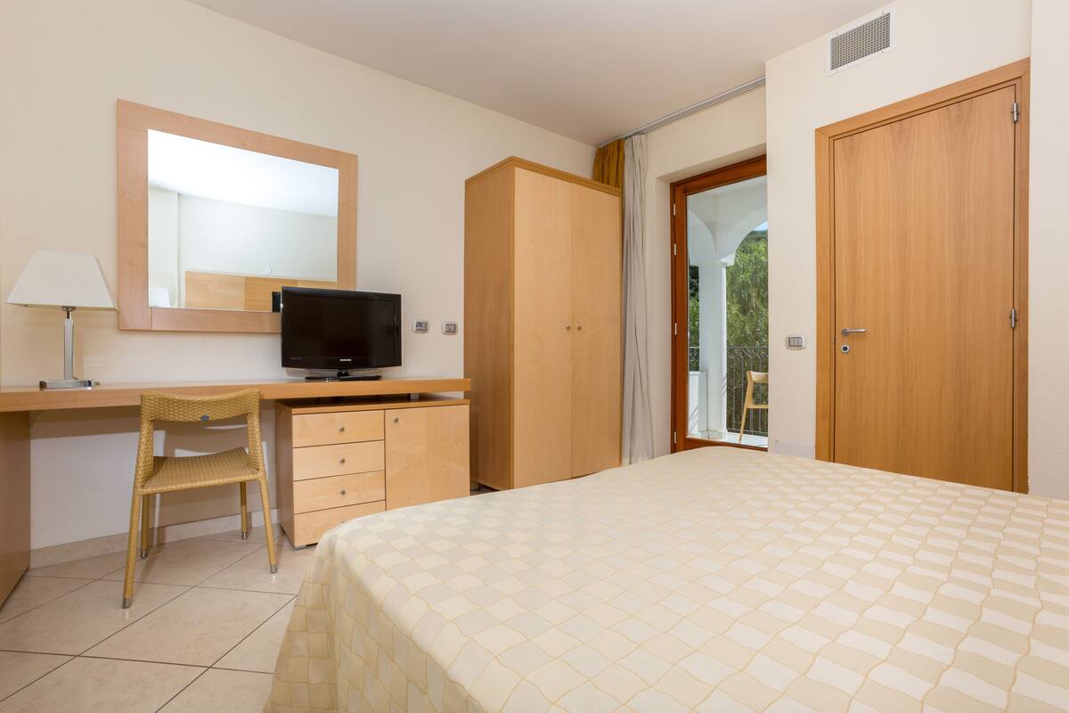 Habitación de hotel con una cama, un escritorio con silla, televisor, armario y puerta que da a un balcón.