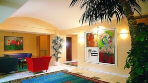 A hotel lobby with a large colorful rug, a seating area with a red chair, a potted plant, and a wall with four framed artworks. - Rokohof (Klagenfurt am Woerthersee)