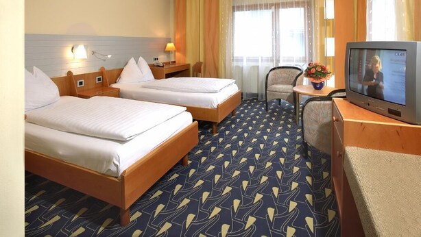 A hotel room with two beds, a desk with a television, a chair, and a small table with a flower. - Rokohof (Klagenfurt am Woerthersee)