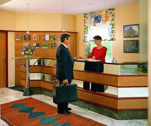A reception desk with a man and woman standing, a plant, and a framed picture on the wall. - Rokohof (Klagenfurt am Woerthersee)