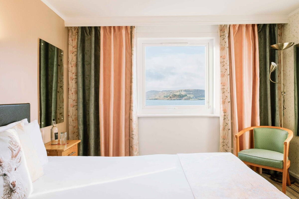 Habitación de hotel con cama, silla, mesita de noche, lámpara y ventana con vista al mar.