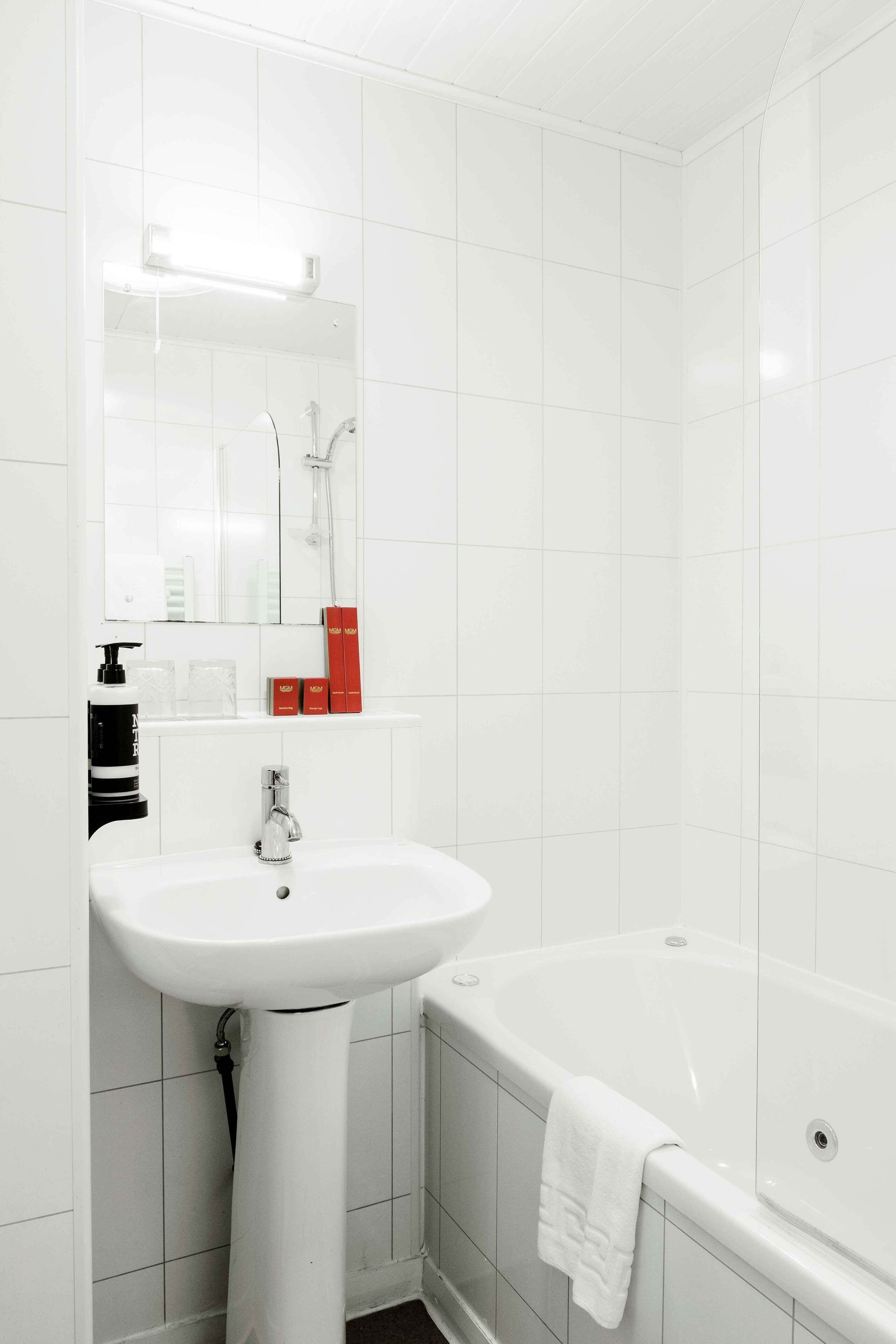A modern bathroom with white tiles, a pedestal sink, a bathtub, and a mirror above the sink.