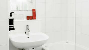 A modern bathroom with white tiles, a pedestal sink, a bathtub, and a mirror above the sink.