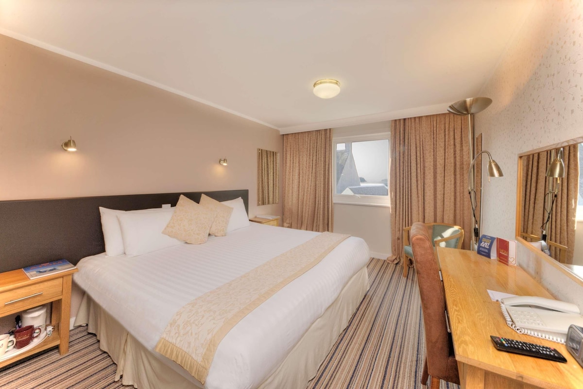 Habitación de hotel con cama, mesitas de noche, un escritorio con silla, teléfono y ventana con cortinas.