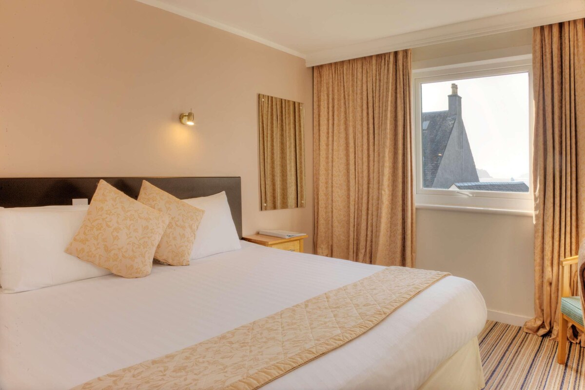 Una habitación de hotel con cama, mesita de noche, ventana con cortinas y vistas a la ciudad.