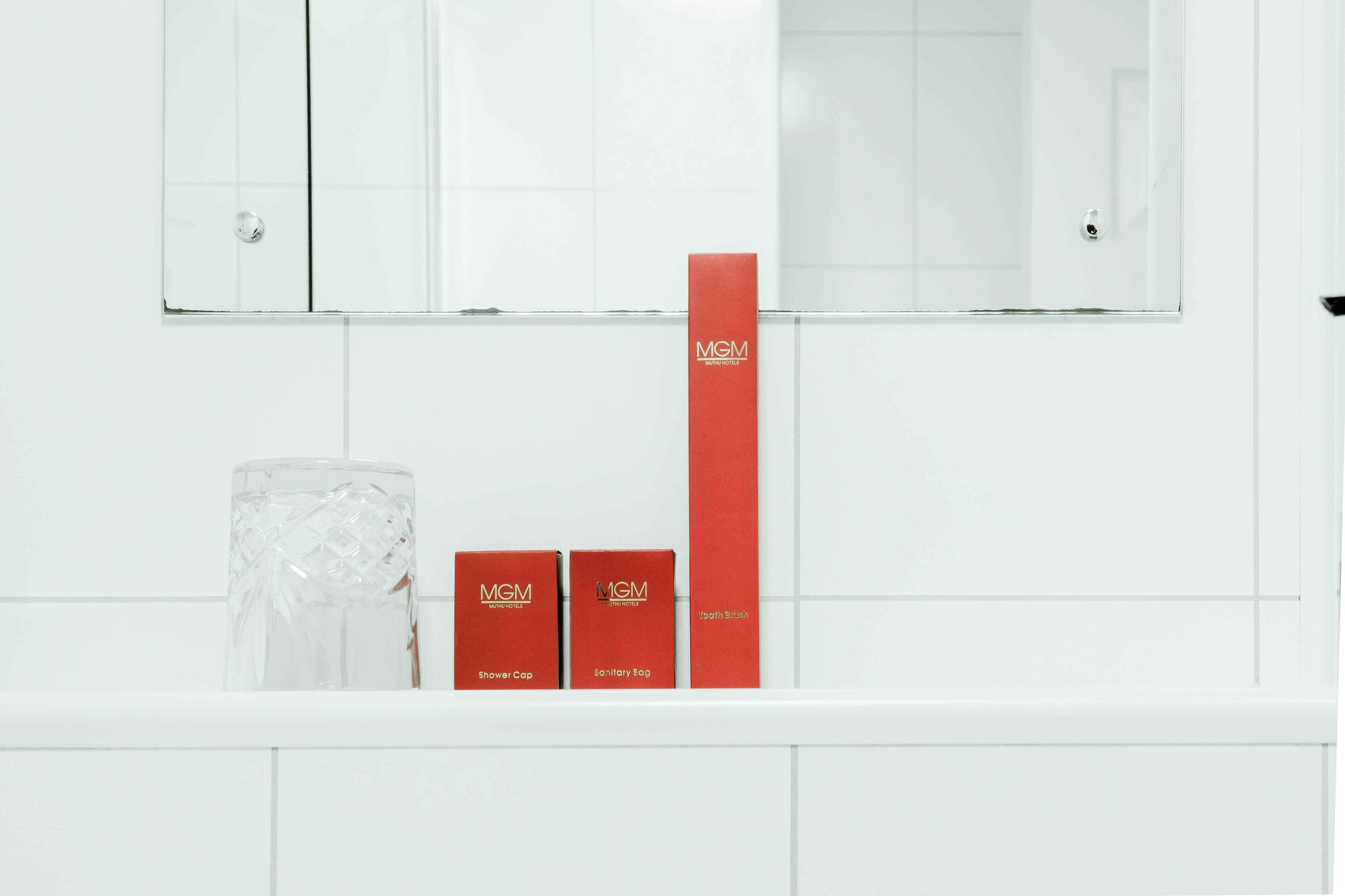 A bathroom with a white tiled wall, a glass shower enclosure, and two red MGM-branded boxes labeled "Shower Cap" and "Antimicrobial Bag".