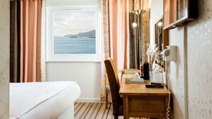 Habitación de hotel con cama, escritorio con lámpara, secador de pelo y vista al mar por una ventana.
