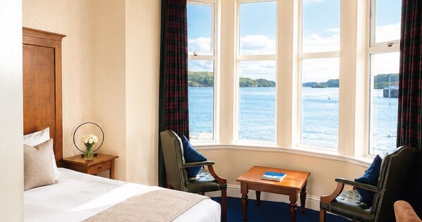 A hotel room with a bed, two chairs, a small table, and a view of a body of water through large windows. - Muthu Queens Hotel (Oban)