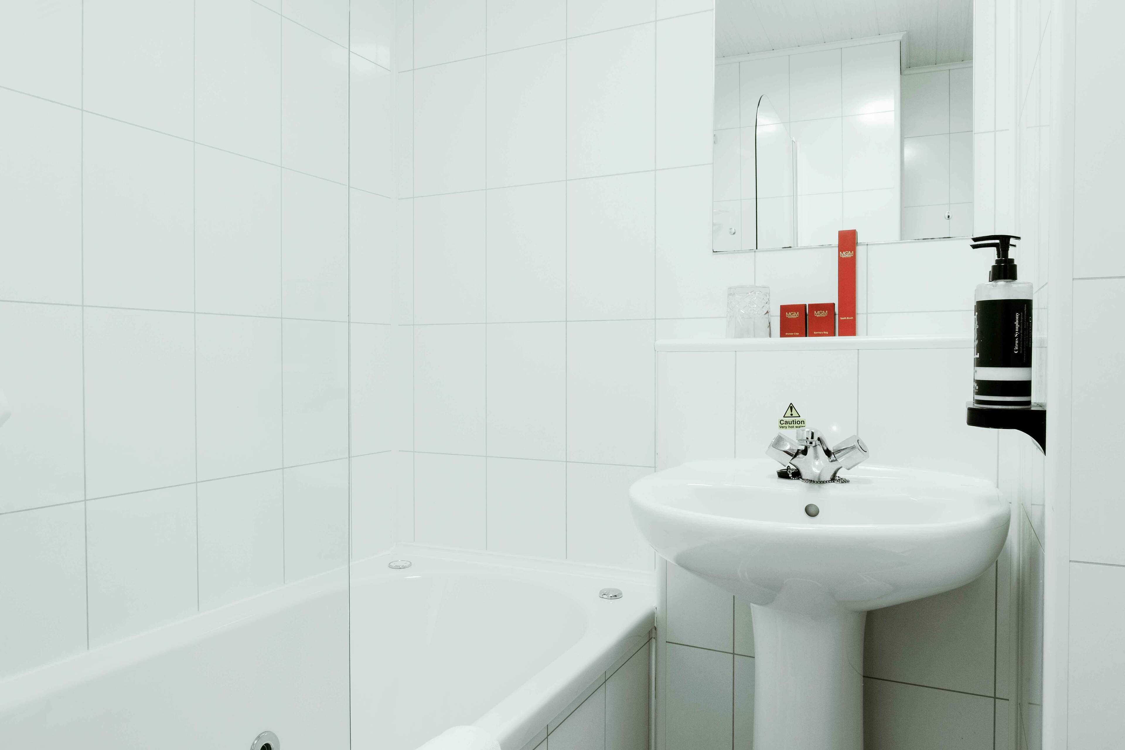 A modern bathroom with white tiles, a white bathtub, a white sink, and a mirror above the sink.