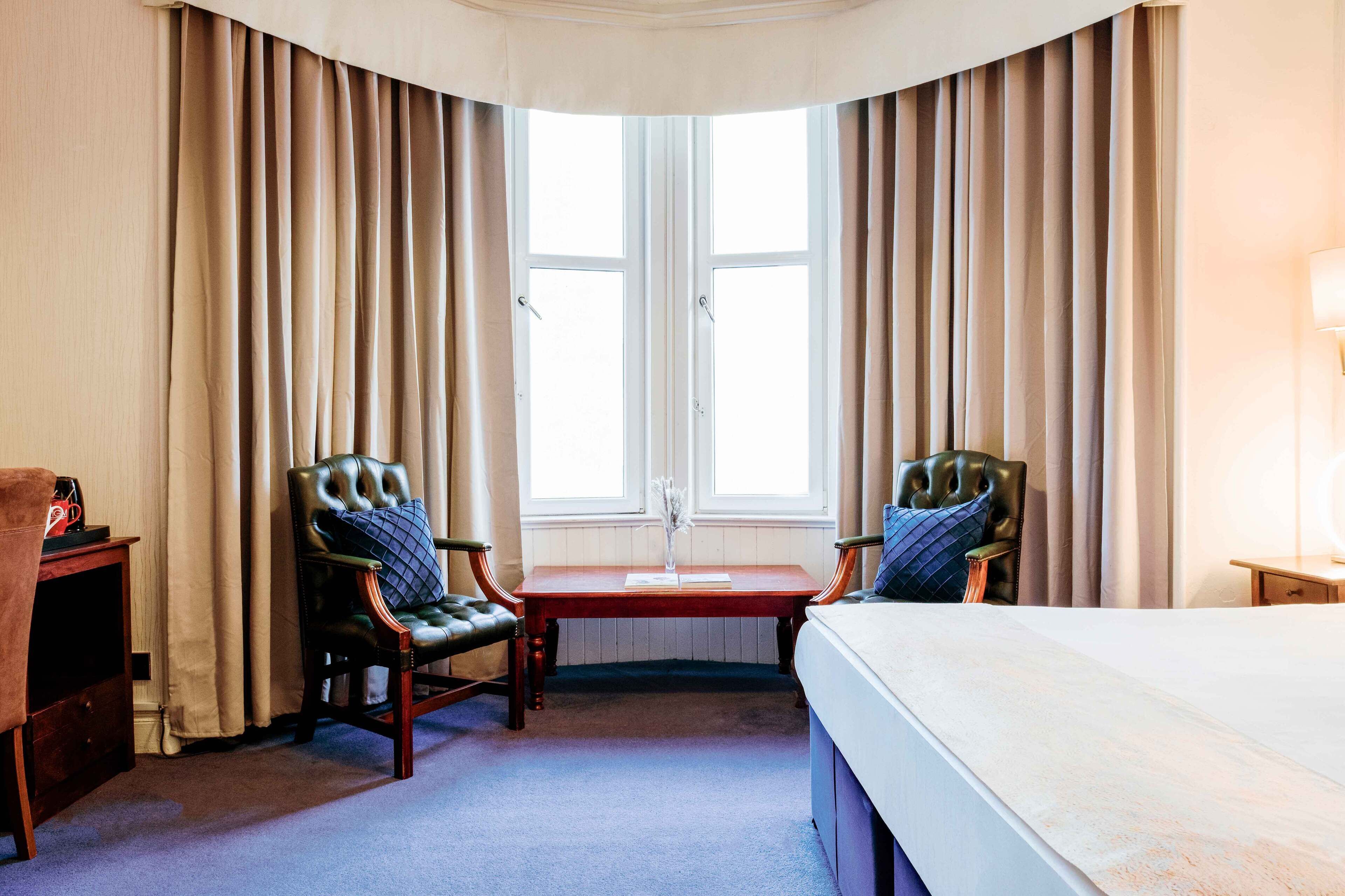Habitación de hotel con una cama grande, dos sillones, una mesita y una ventana con cortinas.