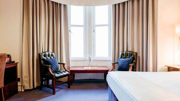Habitación de hotel con una cama grande, dos sillones, una mesita y una ventana con cortinas.