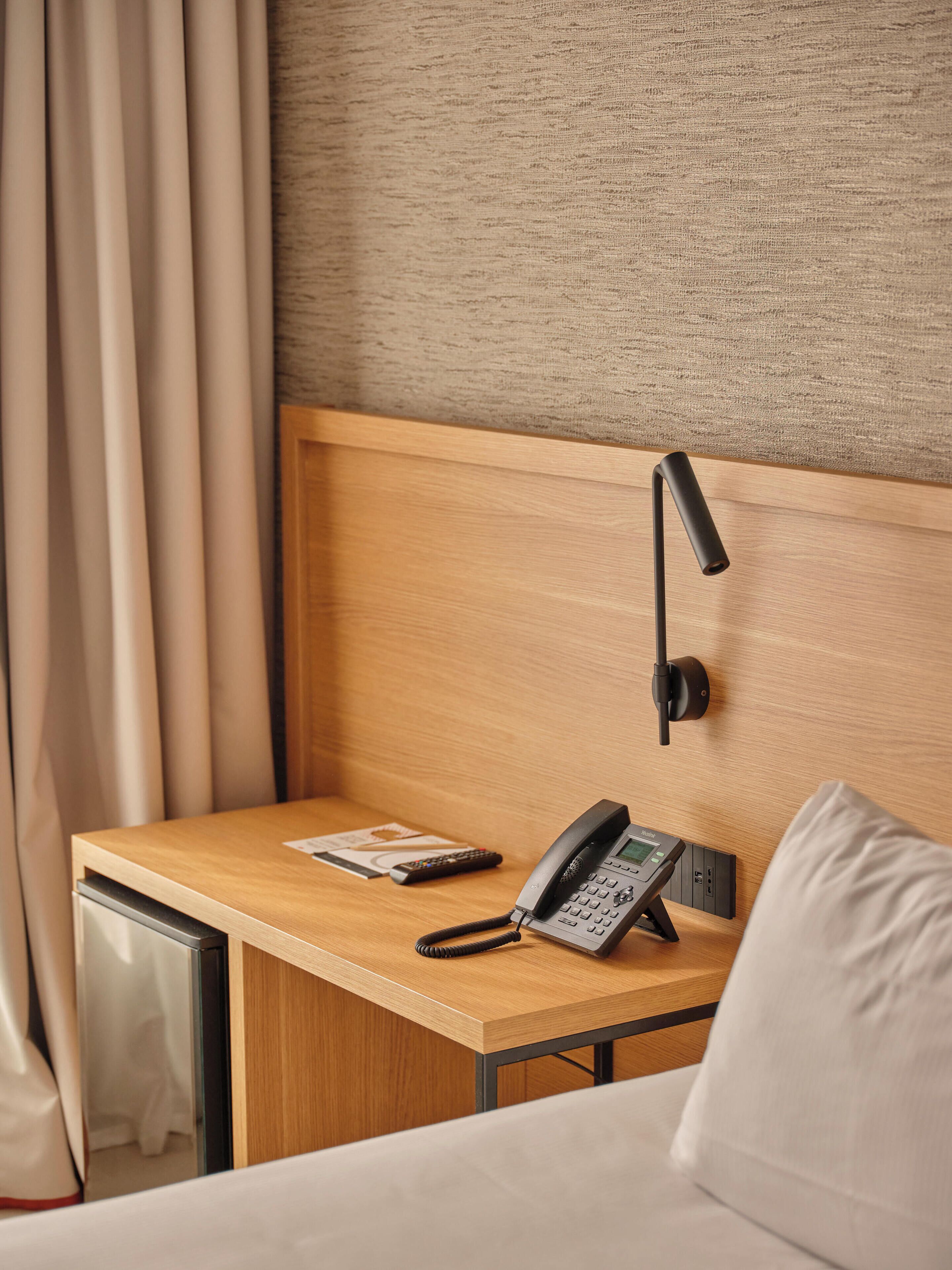 Ein Hotelzimmer mit einem Holztisch, einem Telefon, einer Fernbedienung und einem Bett mit einem weißen Kopfkissen.