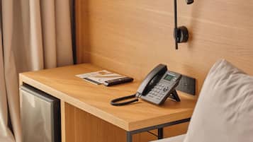 A hotel room with a wooden desk, a telephone, a remote control, and a bed with a white pillow.