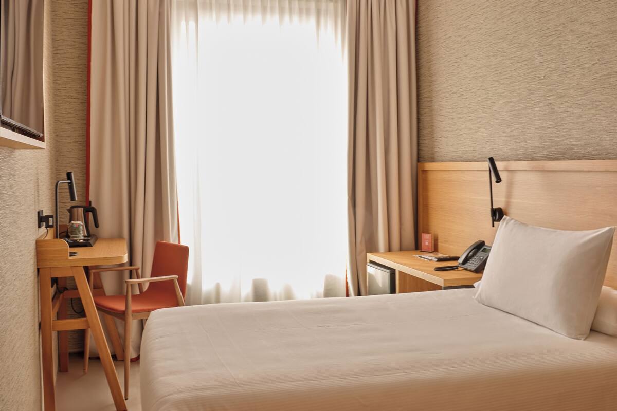 A hotel room with a bed, a desk with a chair, a telephone, and a window with curtains.