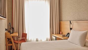 Una habitación de hotel con cama, escritorio, silla, teléfono y ventana con cortinas.