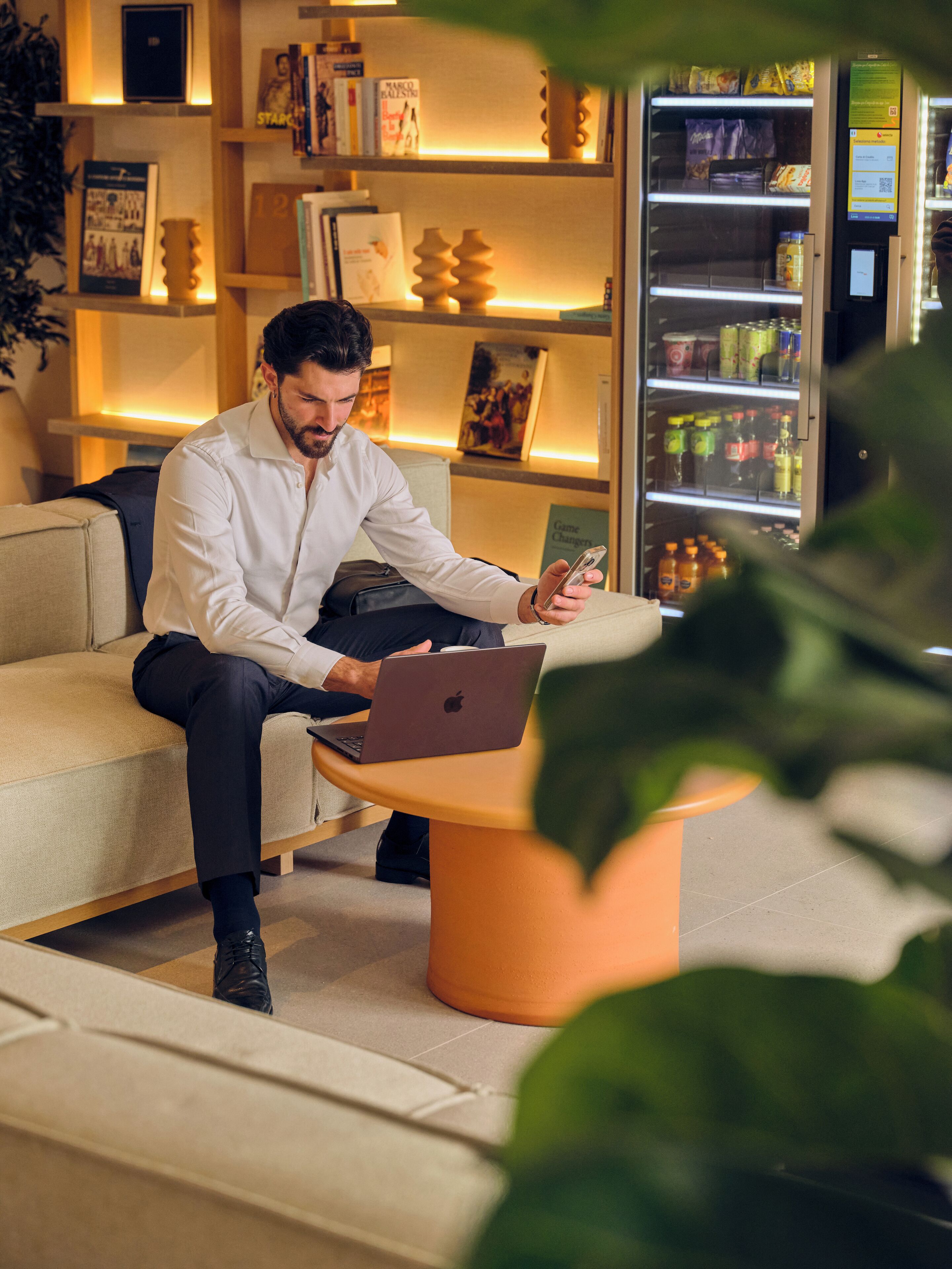 A man sitting on a sofa with a laptop, in a room with a bookshelf and a refrigerator.