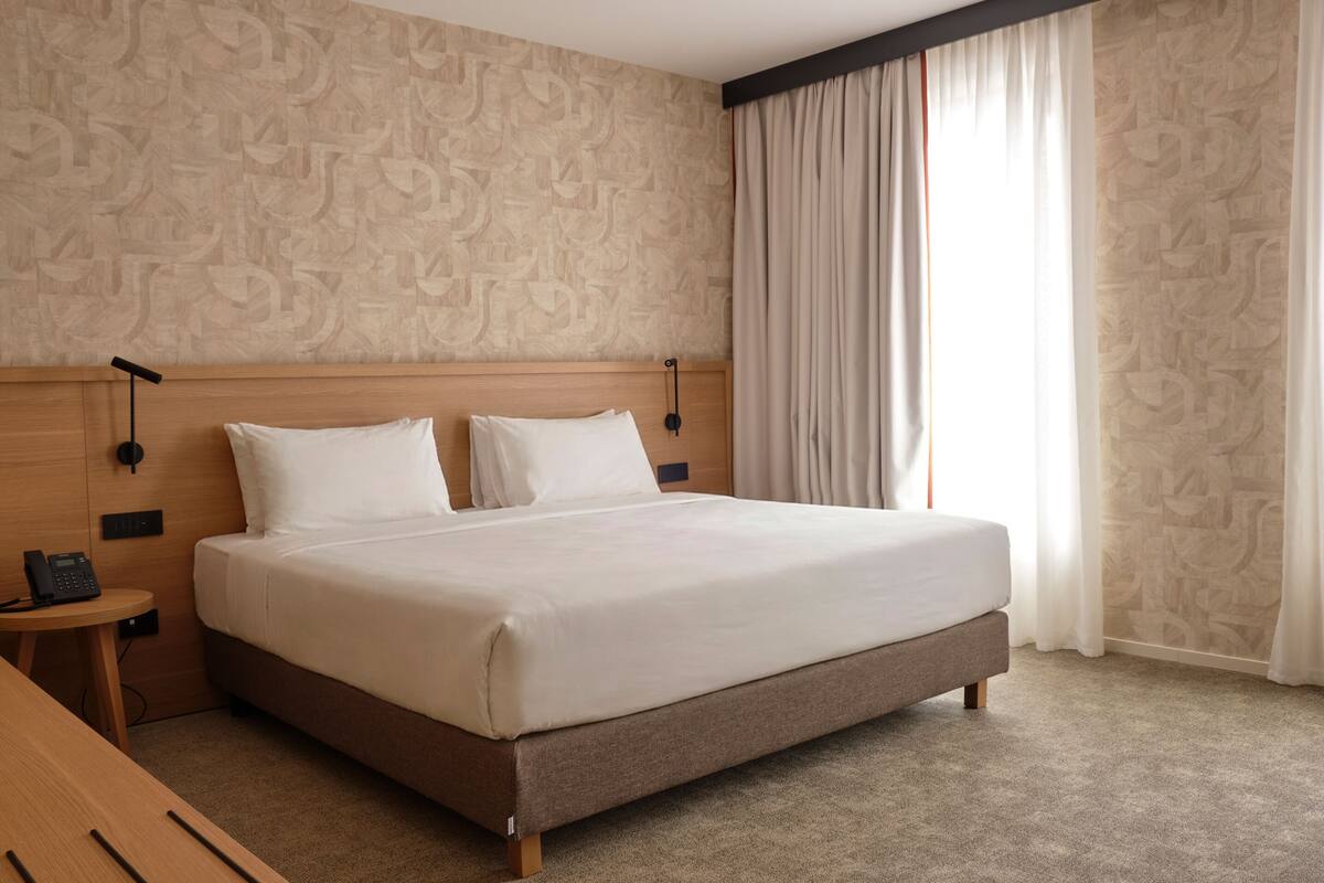 A hotel room with a large bed, a wooden headboard, a bedside table with a phone, and a window with curtains.