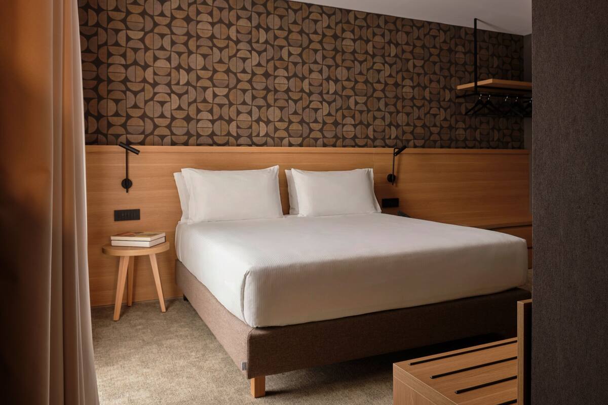 A neatly made bed with a wooden headboard and two pillows, a small wooden nightstand with a book, and a patterned wall behind.