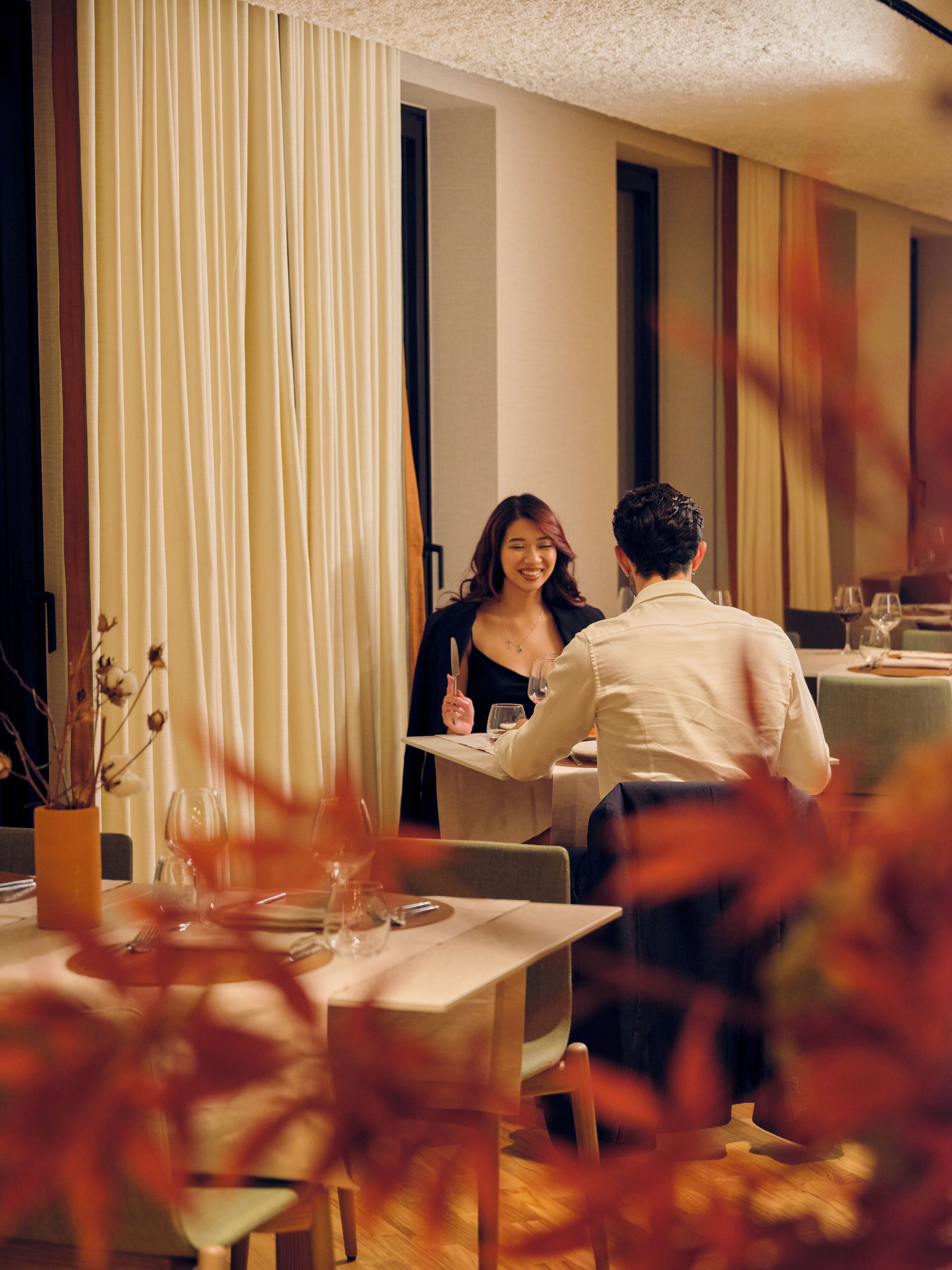 A dining area with a table set for two, a vase with dried flowers, and a curtain in the background.